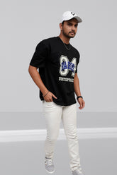 Man wearing a black t-shirt with a logo and text, white pants, and a cap on a gray background