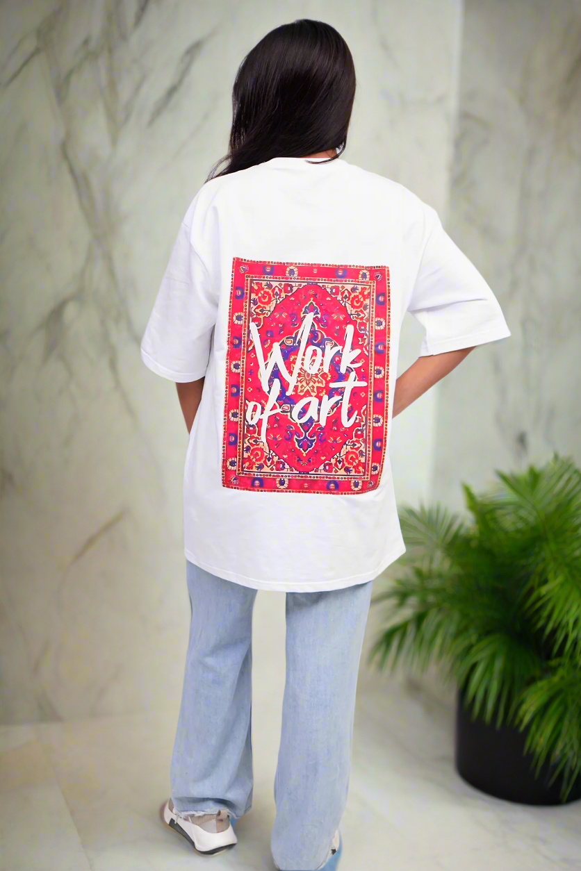 Person wearing a white t-shirt with a colorful graphic on the back, standing in a room with a plant and marble wall.