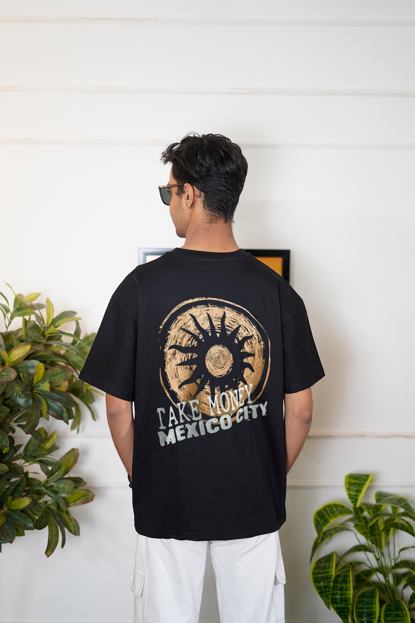 Person wearing a black t-shirt with 'Take Money Mexico City' design, standing indoors with plants in the background.