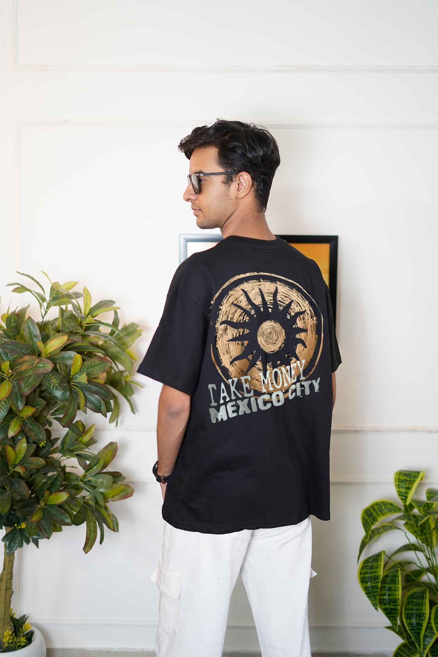 Person wearing a black t-shirt with a design and text, standing indoors with plants in the background.
