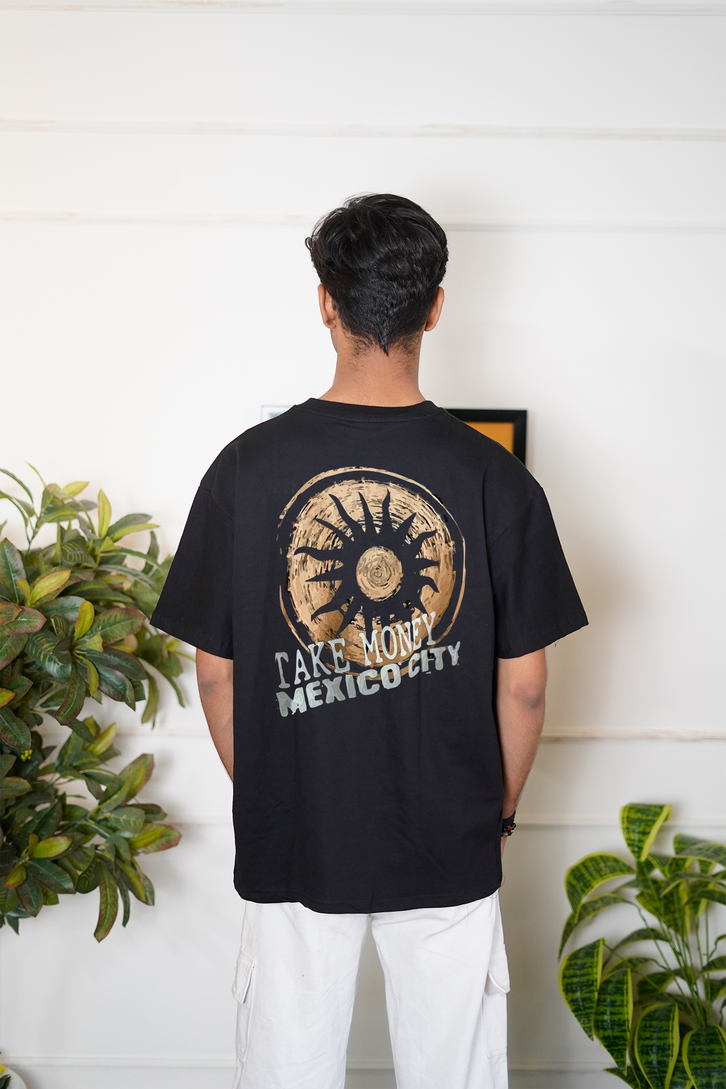 Person wearing a black t-shirt with a design and text, standing in front of a white wall with plants.