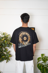 Person wearing a black t-shirt with a design and text, standing in front of a white wall with plants.