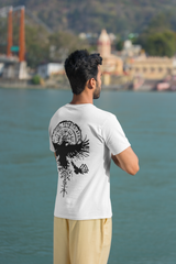 Man wearing a white t-shirt with a black design, standing by a body of water with a scenic background.