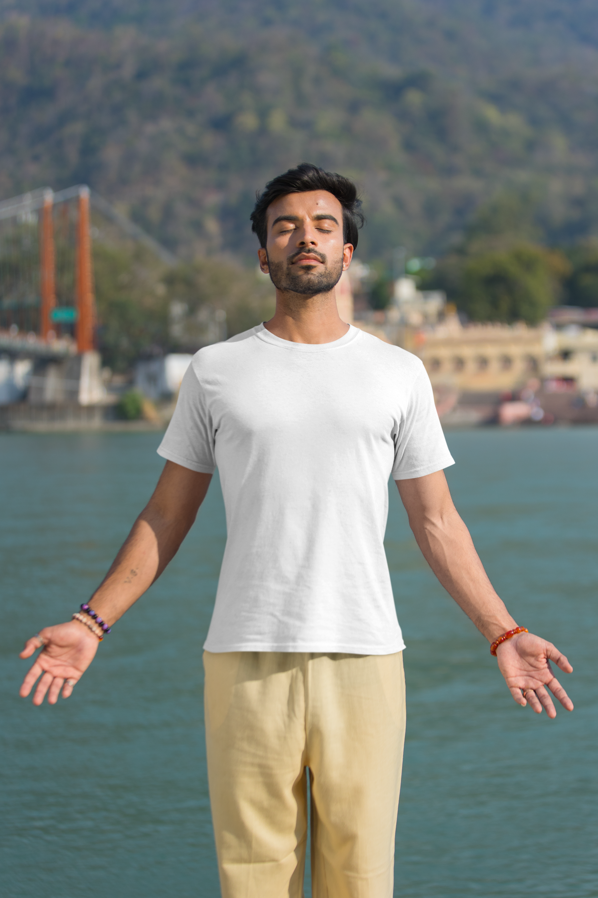 Man in a white t-shirt and beige pants standing by a body of water with mountains in the background