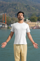 Man in a white t-shirt and beige pants standing by a body of water with mountains in the background