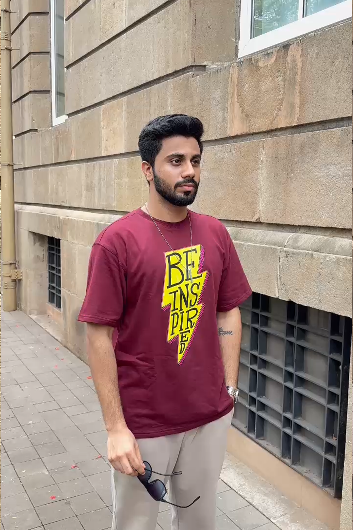 Man wearing a maroon t-shirt with a yellow graphic design standing against a stone wall.