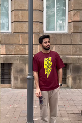 Man wearing a maroon t-shirt with yellow text standing against a brick wall.