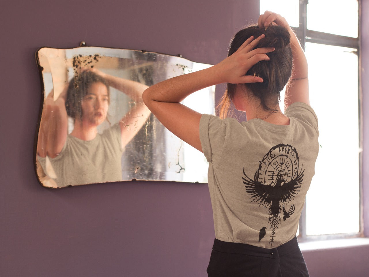 Person adjusting hair in front of a mirror with a decorative frame, wearing a t-shirt with a black design.