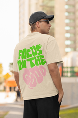 Person wearing a beige t-shirt with green and pink text outdoors  Oversized T-Shirt From Kapda Kulture.