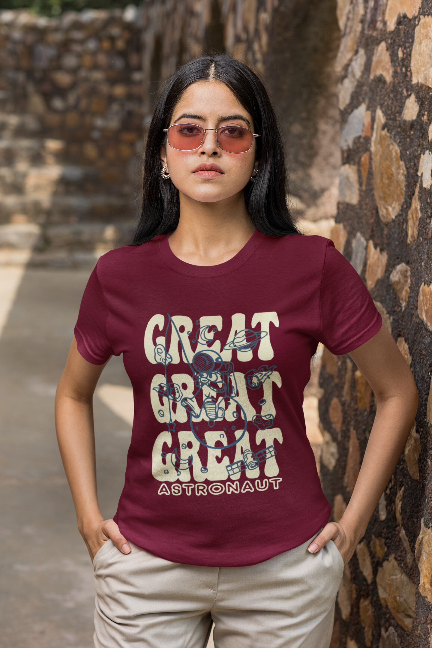 Woman wearing a maroon t-shirt with 'GREAT GREAT GREAT ASTRONAUT' text and sunglasses, standing against a stone wall.