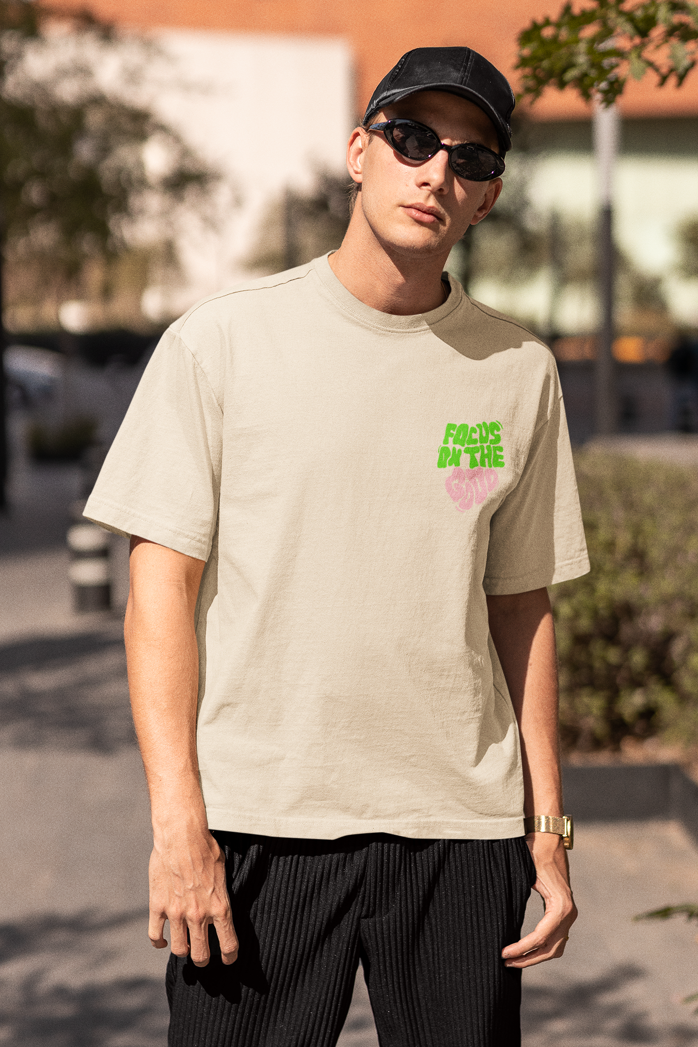 Person wearing a beige t-shirt with green text, black cap, and sunglasses outdoors  Oversized T-Shirt From Kapda Kulture.