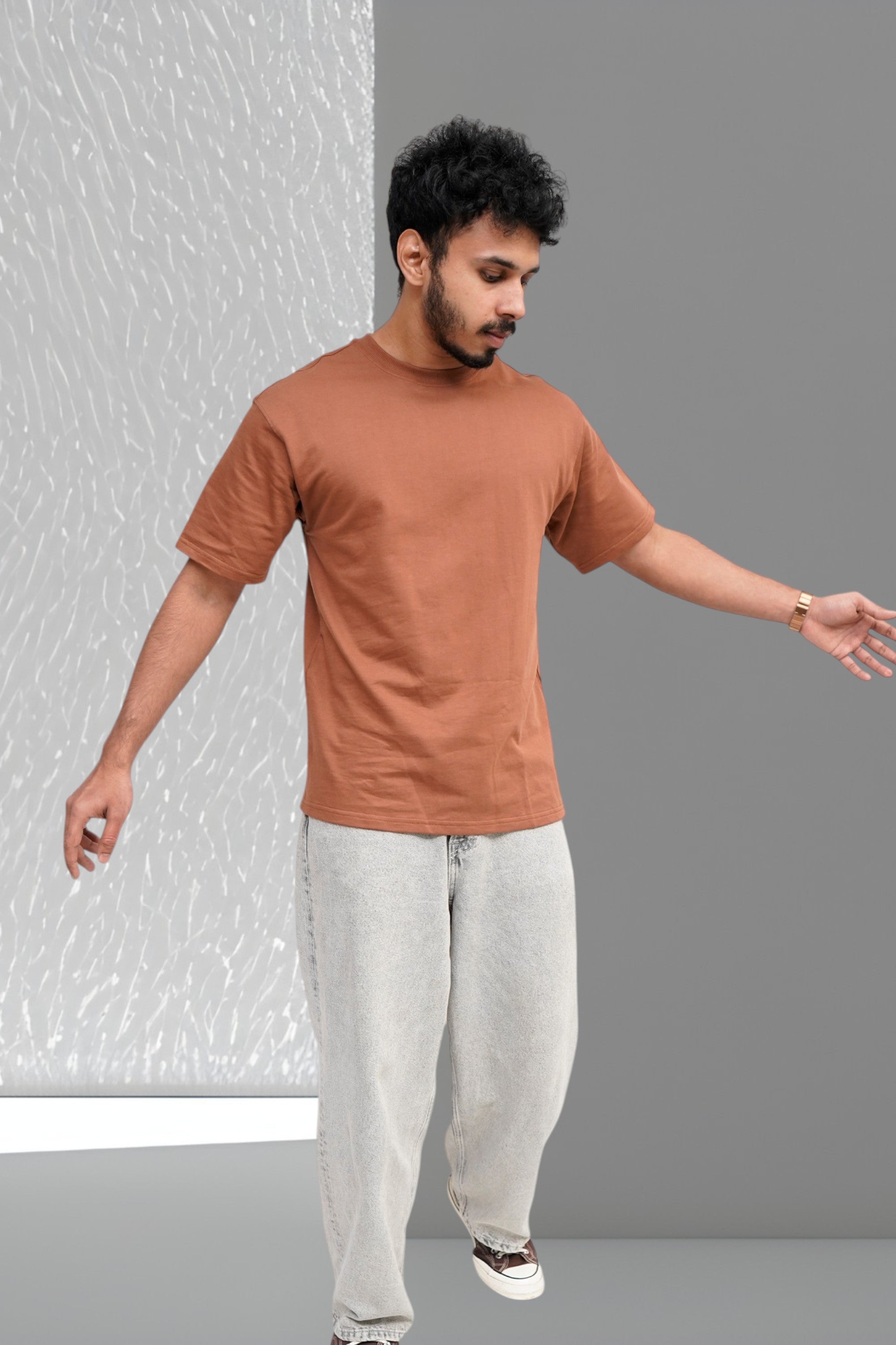 Man wearing a brown t-shirt and light gray pants standing against a plain background